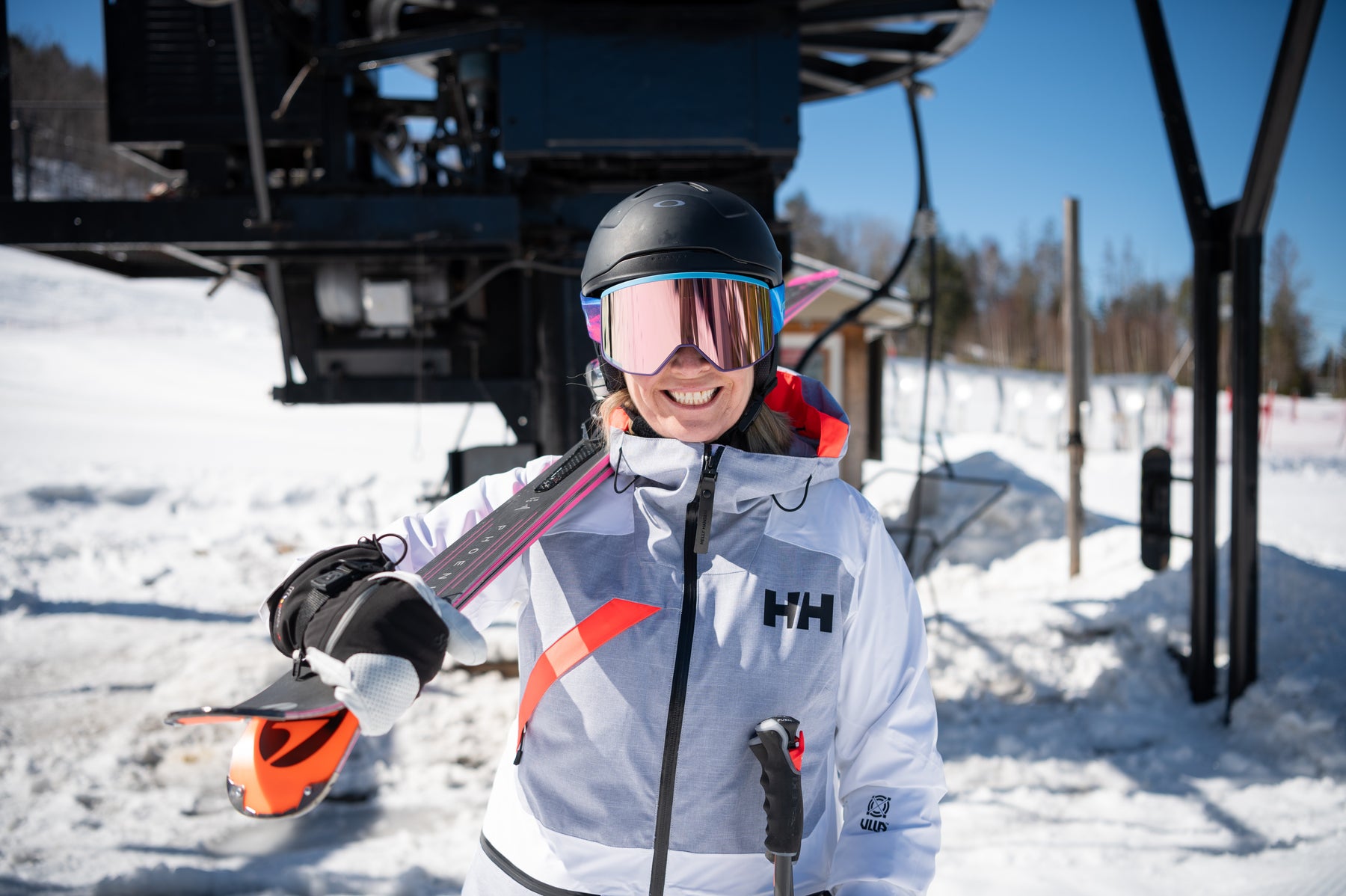 Une skieuse souriante, skis sur l'épaule, devant une remontée mécanique.